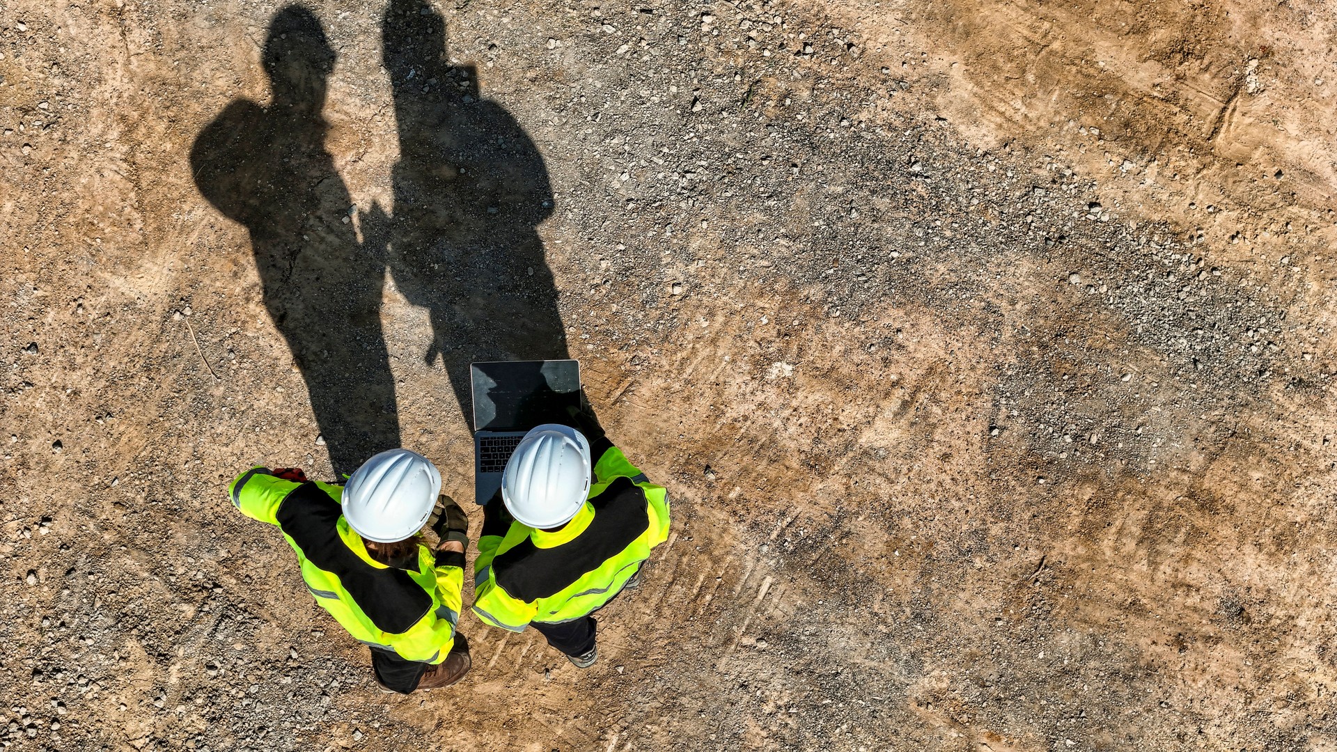 Top-down view of two engineers wearing safety vests and helmets using a laptop on a construction site, with their long shadows cast on the ground.